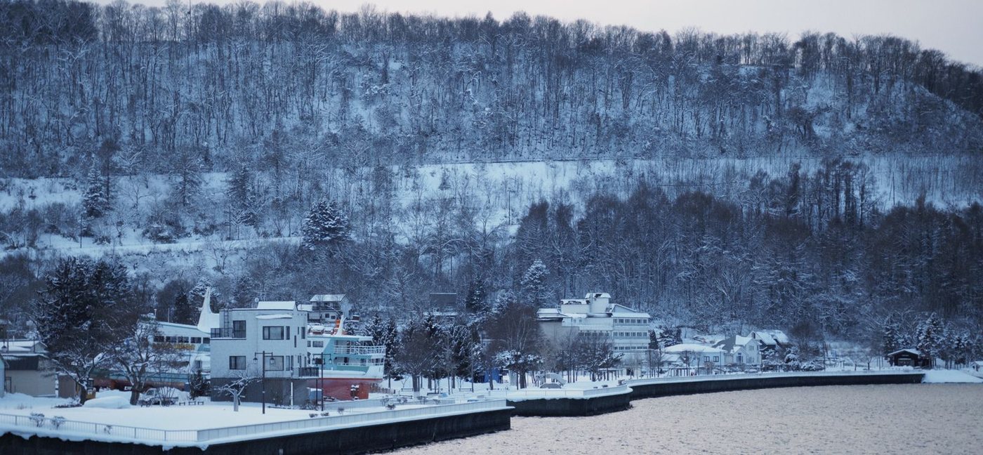 雪の積もった冬の湖岸と、山の斜面に並ぶ建物